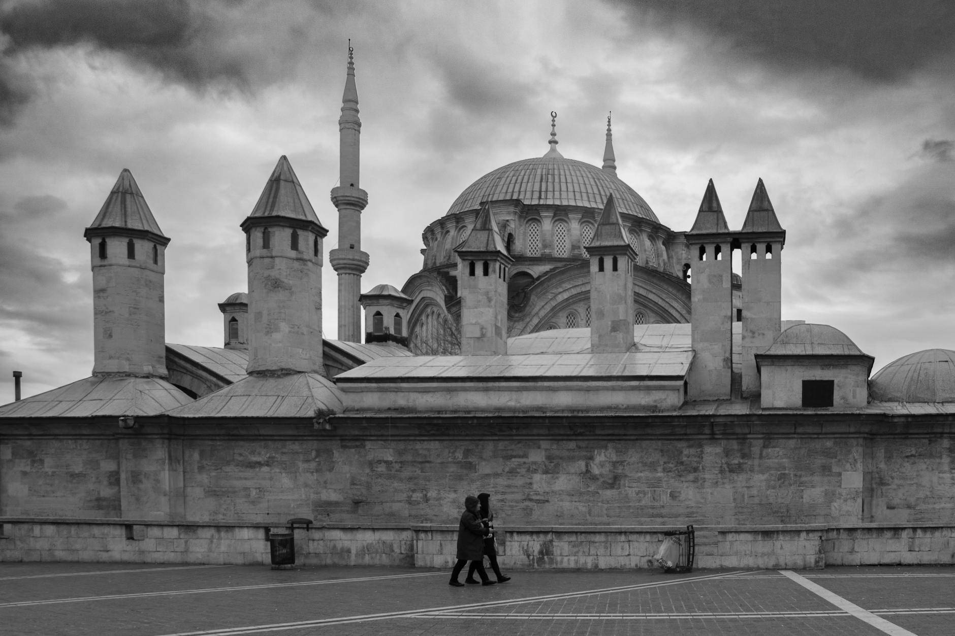 A dramatic, high-contrast black and white photograph showing the unique architecture of the Arap Mosque (or Arap Cami) in Karaköy, Istanbul. The image features the mosque's distinctive stone turrets and a large central dome rising above a substantial stone wall, illustrating why this site is described as 'A Gothic Secret in the Heart of Karaköy: Why I Love the Unique Arap Mosque.' Two figures walk along the paved foreground under a brooding, cloudy sky.