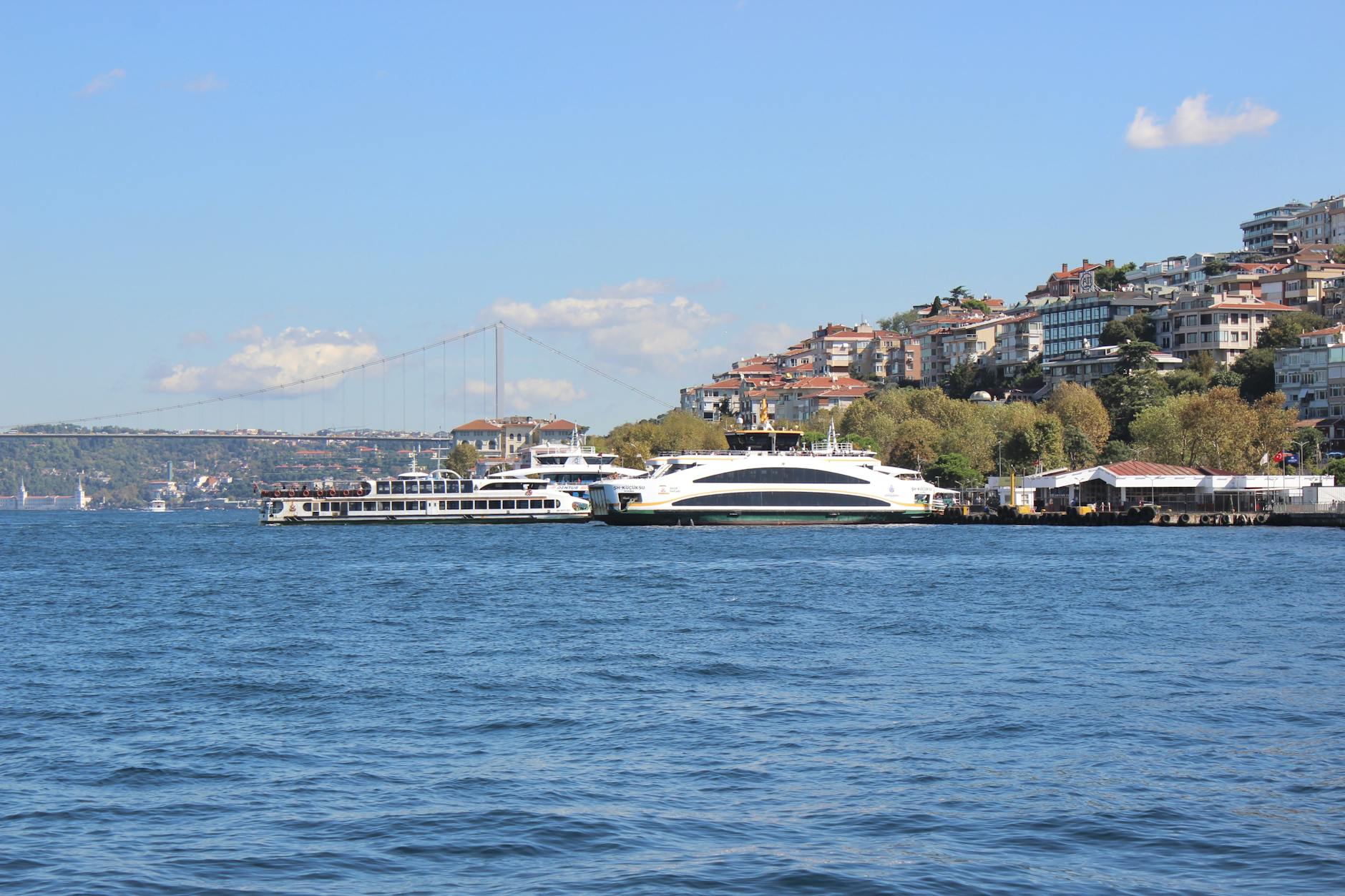 Two ferries docked near a shoreline with densely packed residential buildings and trees climbing the hill, with the Bosphorus Bridge visible in the distance. This captures the scenery of Istanbul tourism.