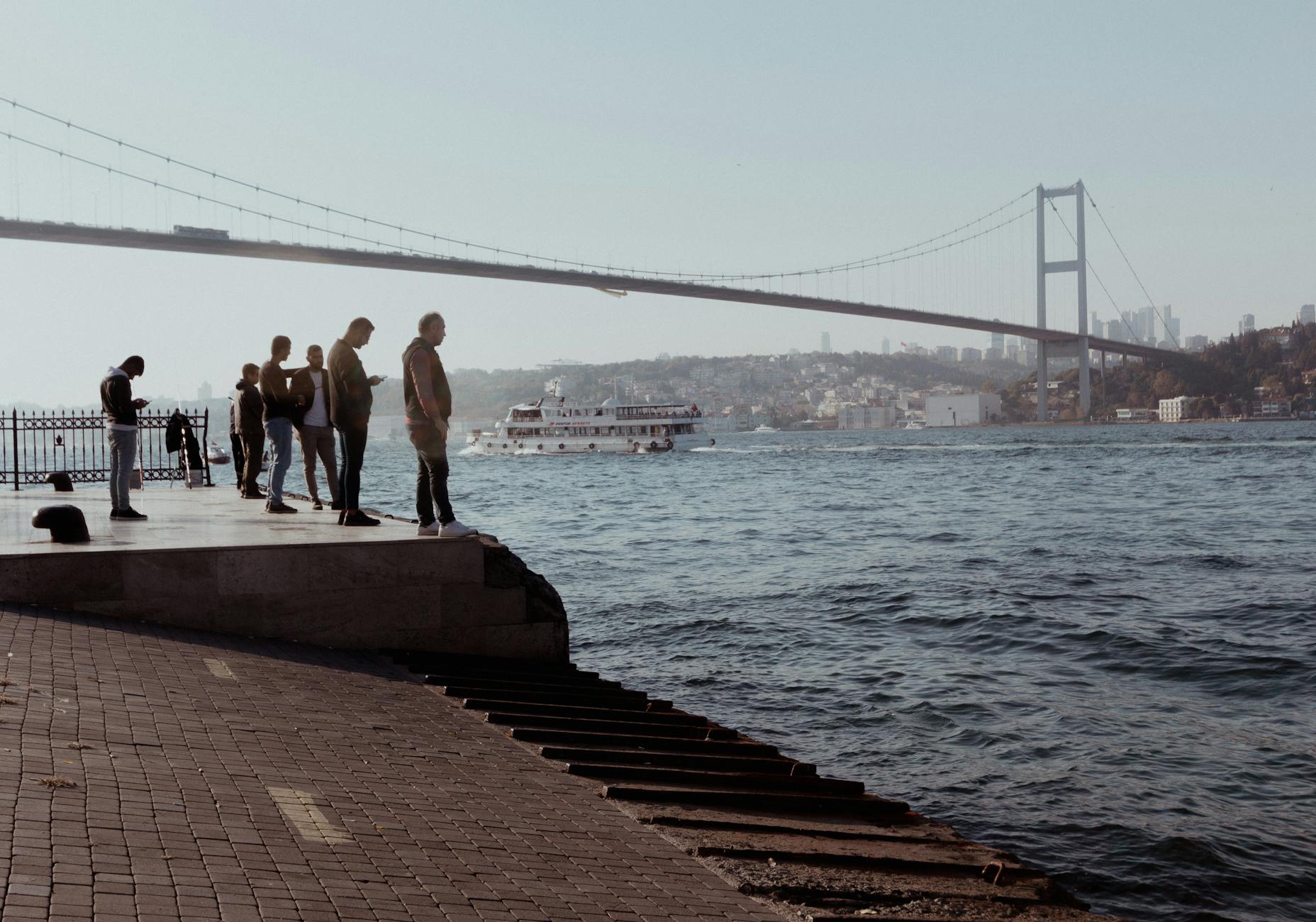 People stand on a stone waterfront looking at a ferry boat on the Bosphorus.