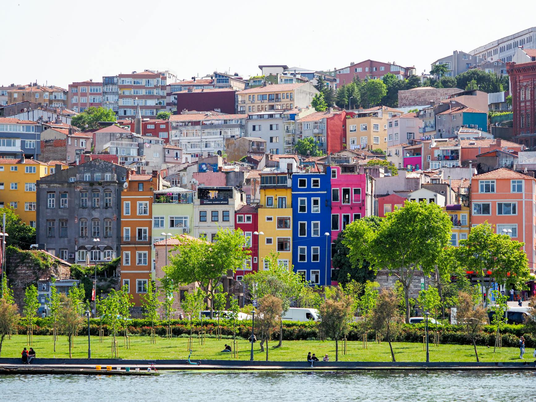 Vibrant, colorful historic buildings of Istanbul rising behind a green waterfront park area by the water, suggesting scenic views near Balat or Fener.