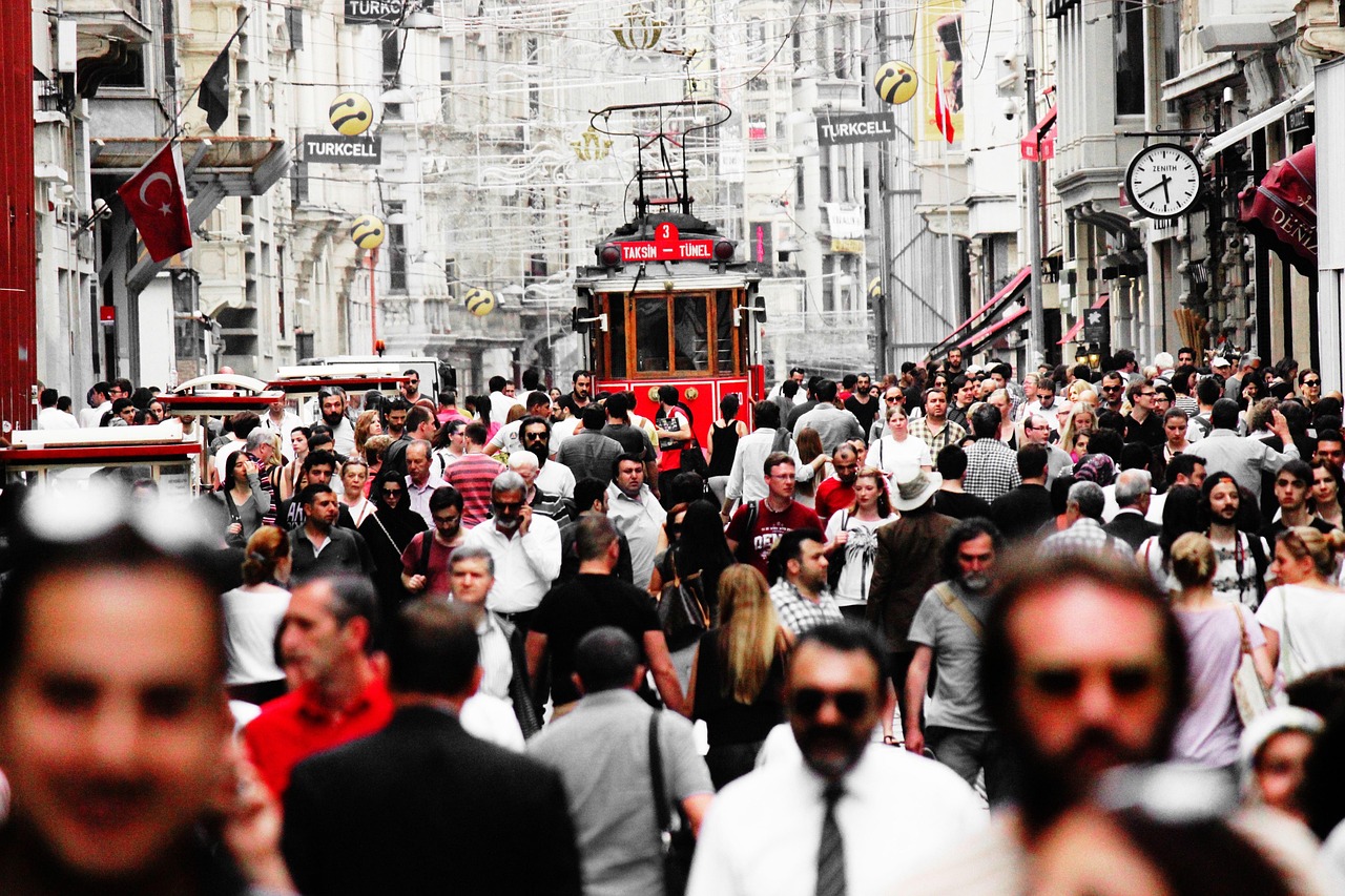 A high-density crowd of people fills a narrow, historic street in Istanbul, evoking the feeling of 'The frantic sweaty madness of my walk through Mahmutpaşa and Tahtakale.' In the center background, the iconic red T3 Taksim-Tünel heritage tram is visible, surrounded by shoppers and pedestrians beneath hanging signs, including one for Turkcell and an outdoor clock face. The image uses selective color to highlight the red tram and some clothing against muted building tones.
