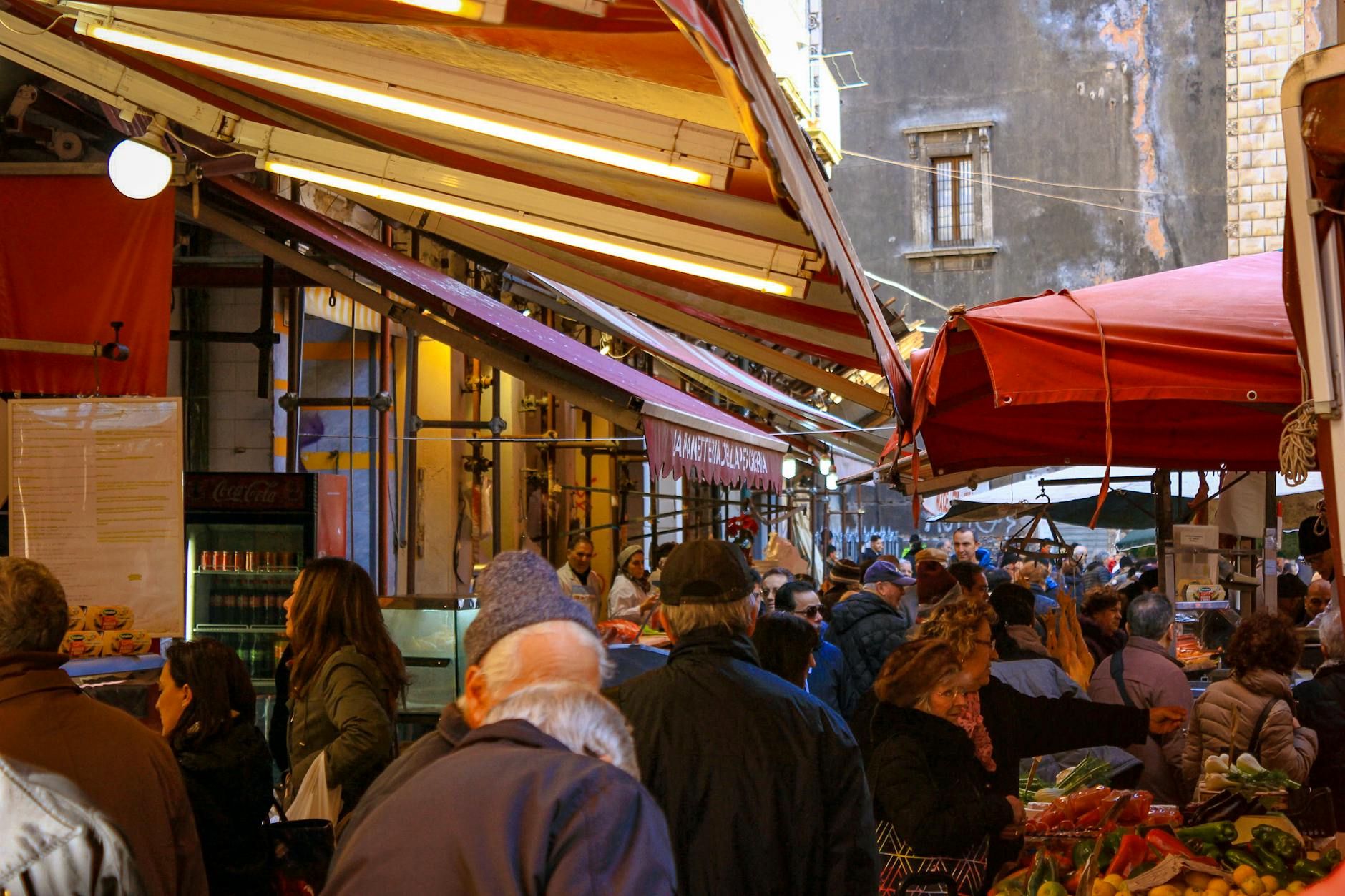 Shoppers navigate a crowded outdoor street market filled with fresh produce under colorful awnings.