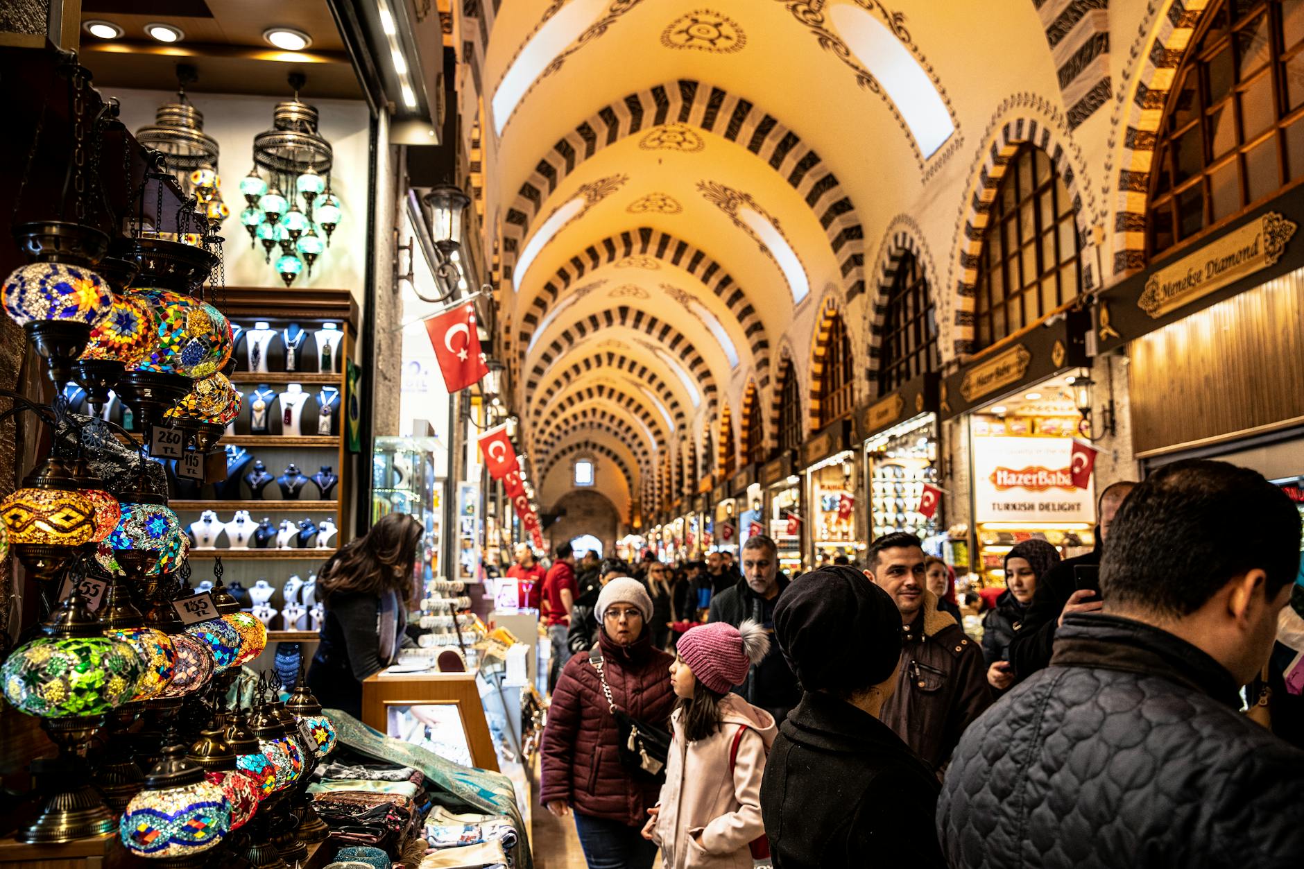 Colorful mosaic Turkish lamps displayed inside the historic Grand Bazaar in Istanbul with shoppers browsing the stalls.
