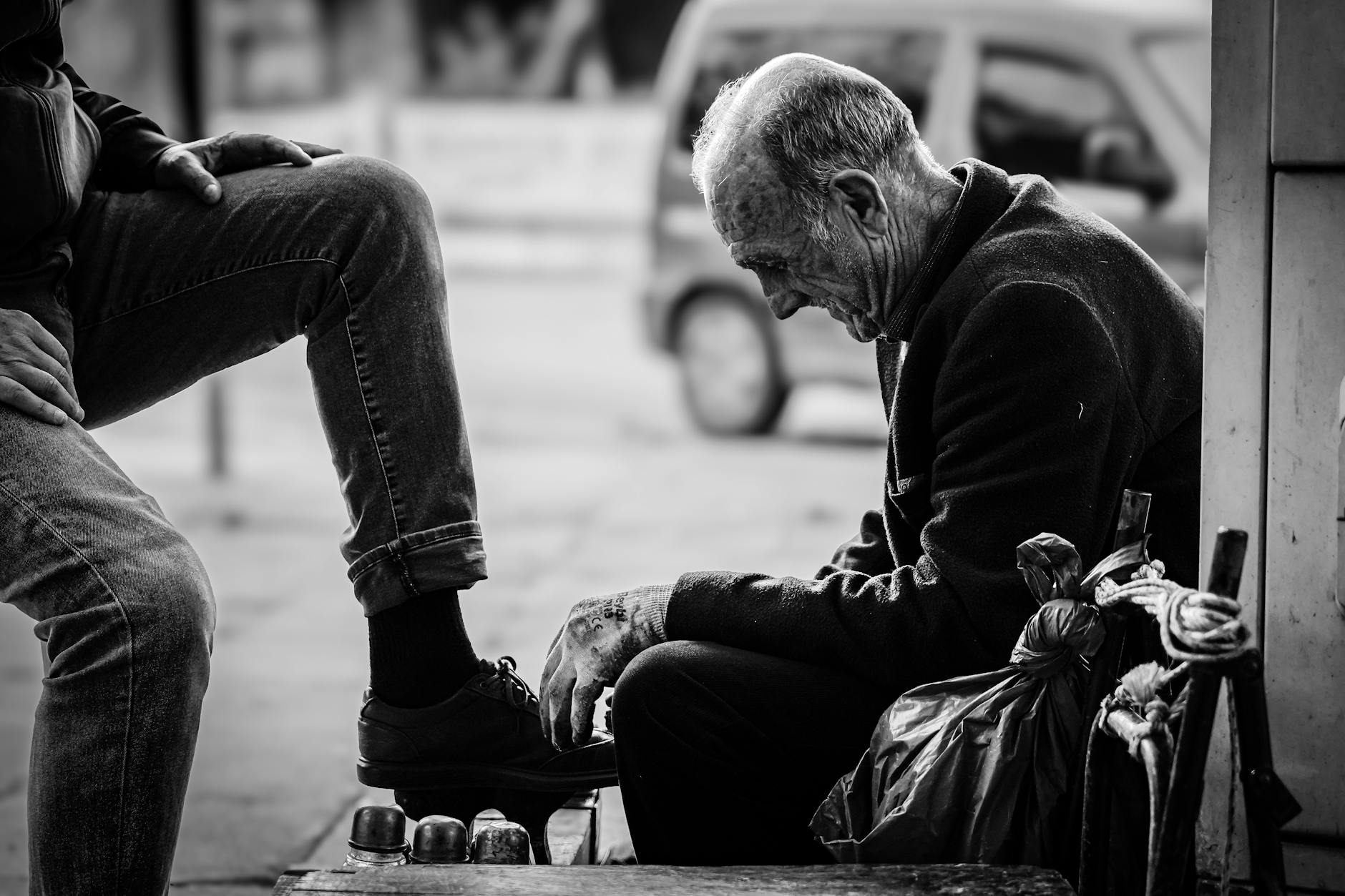 A poignant, black and white street photograph captures the intimate scene of an elderly shoeshine man diligently working on a customer's shoe, set against the blurred backdrop of a busy urban area, typical of the dense atmosphere one experiences during 'The frantic sweaty madness of my walk through Mahmutpaşa and Tahtakale' in Istanbul.