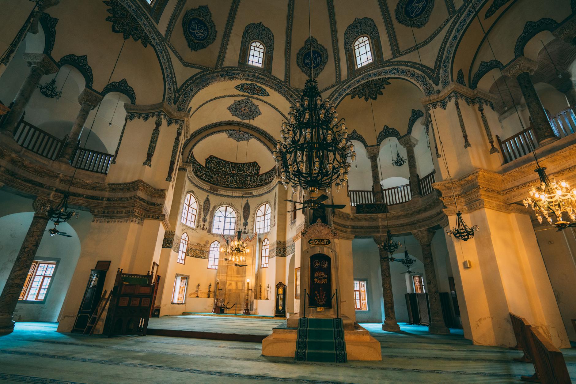 The grand, dimly lit interior of the Little Hagia Sophia (Küçük Ayasofya Camii) in Istanbul, featuring high ceilings, ornate Islamic calligraphy and blue tilework, large chandeliers, and an elevated minbar area. This peaceful sanctuary offers a perfect alternative for those who are done with the Sultanahmet crowds so they go to Little Hagia Sophia instead.