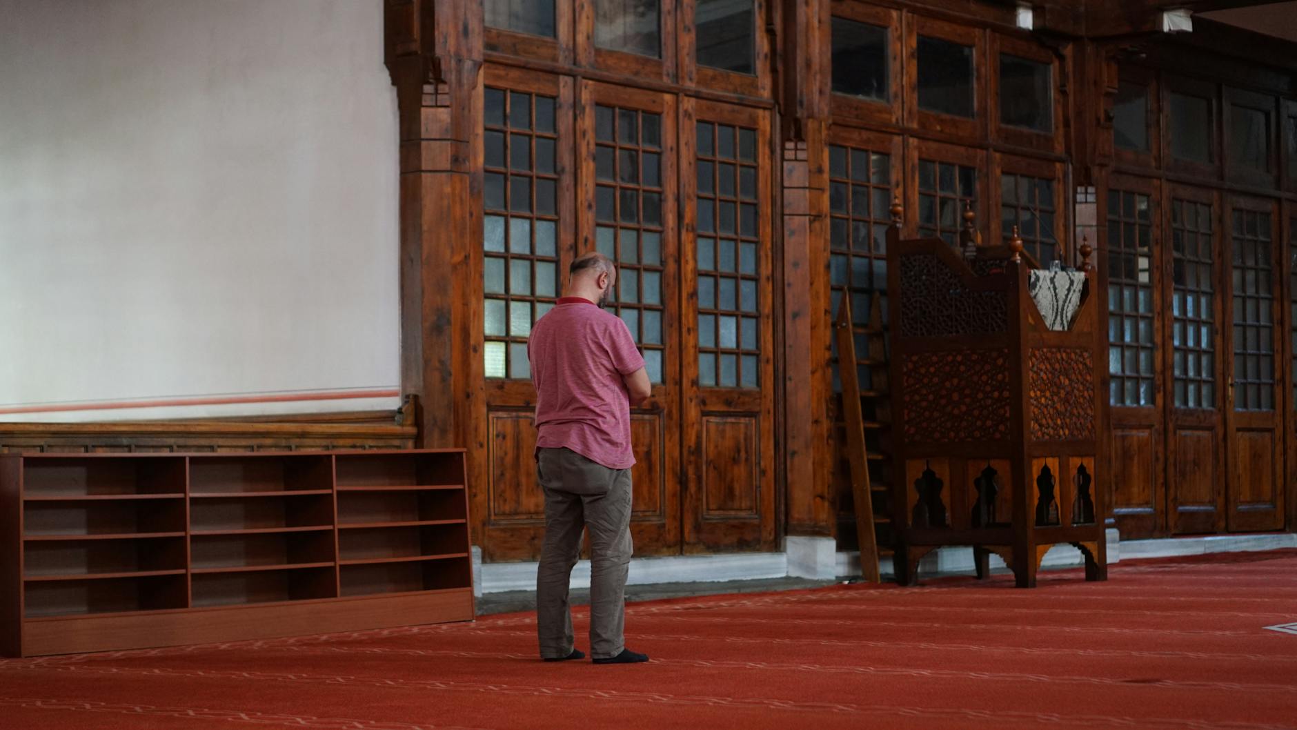 Interior view of a prayer hall featuring rich wooden paneling and doors, with a man standing facing the Qibla wall, possibly inside the unique Arap Mosque that is described as 'A Gothic Secret in the Heart of Karaköy: Why I Love the Unique Arap Mosque'.
