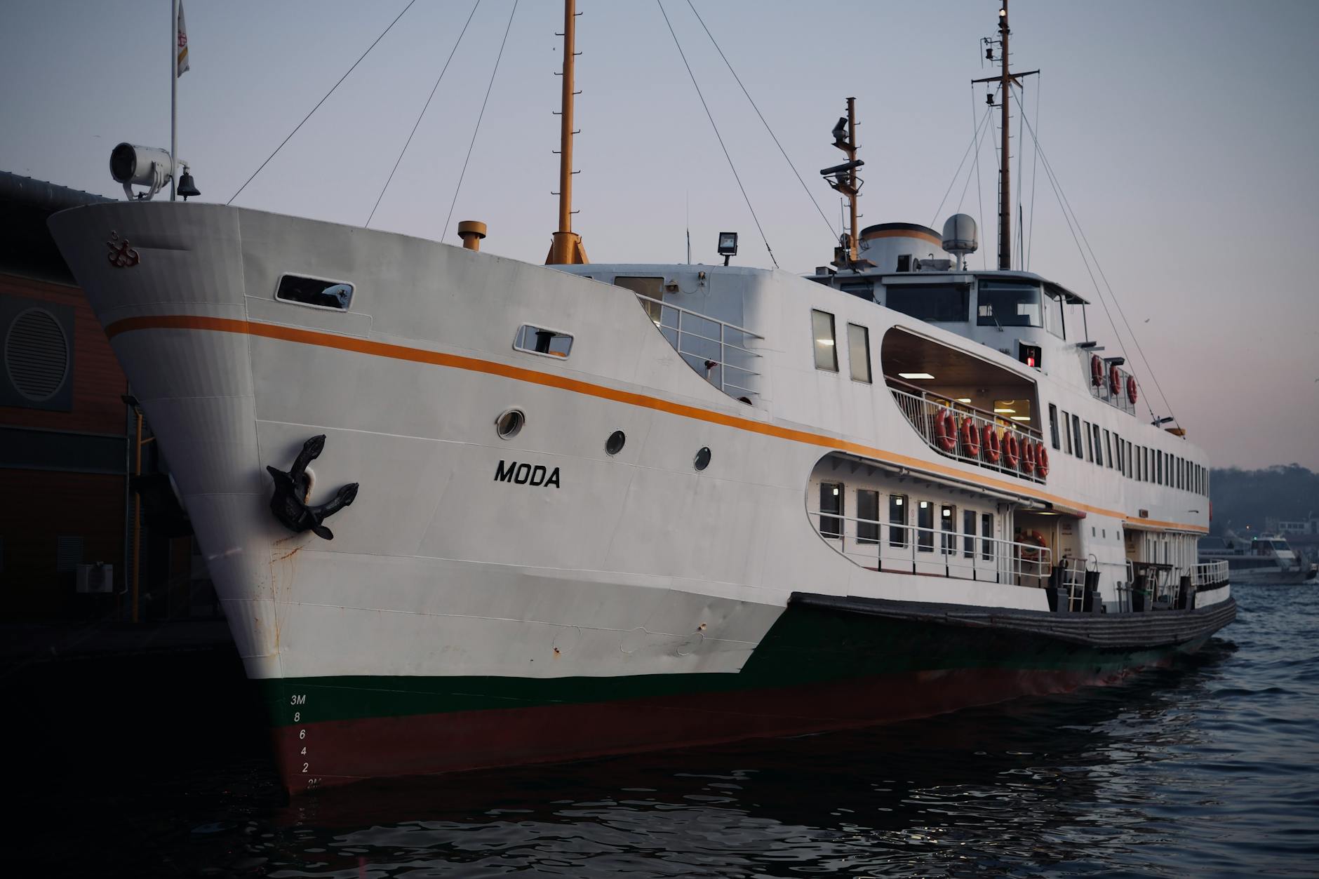 The bow of the white Istanbul ferry named 'Moda' docked at the pier, showcasing its anchor and draft markings, indicative of local sea travel.