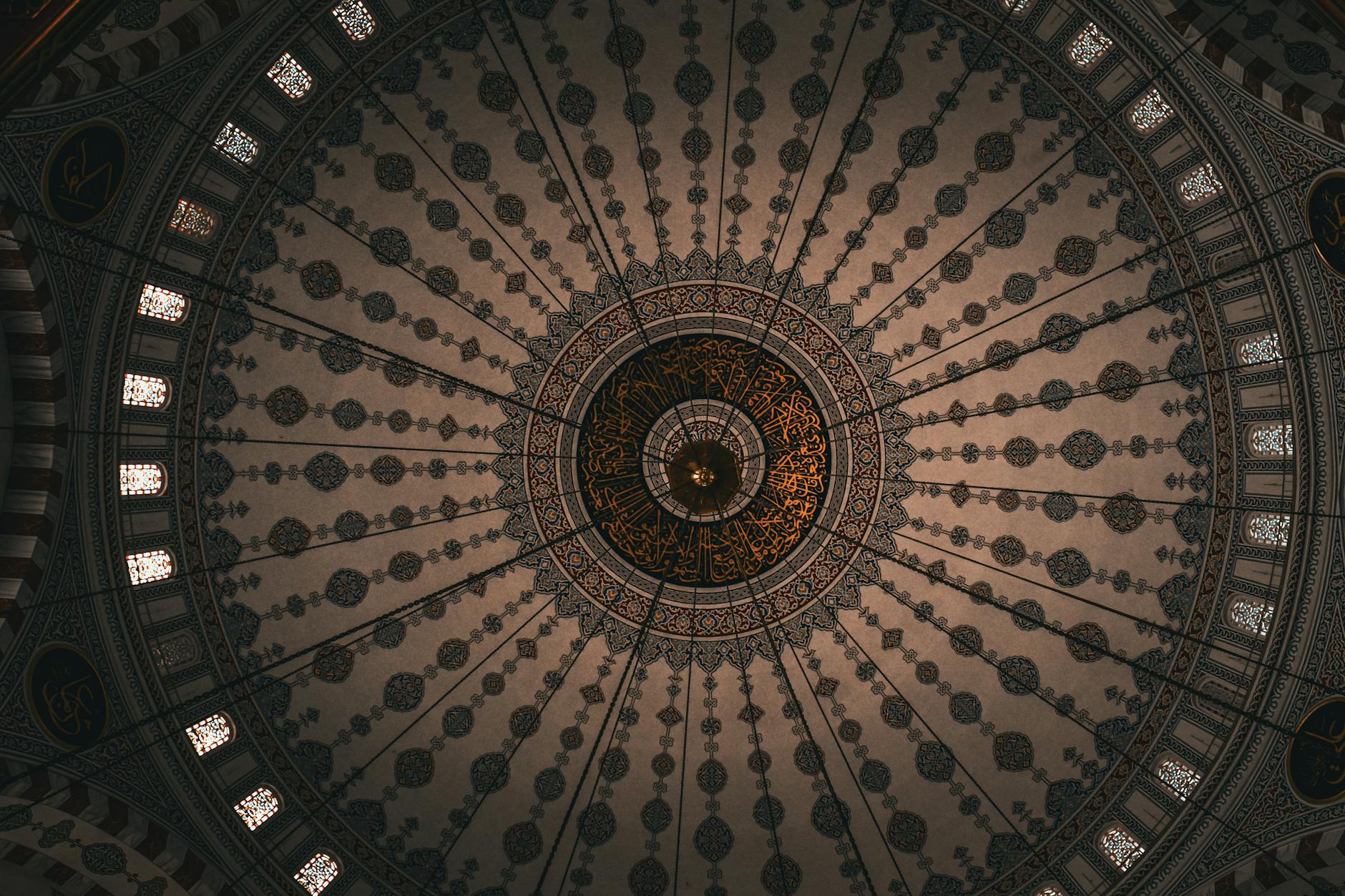 An interior view looking directly up at the highly decorated dome ceiling of an Ottoman mosque, showcasing intricate geometric patterns, radial chain supports, and detailed Arabic calligraphy illuminated in gold at the center. The ambient light filtering through the stained glass windows around the drum highlights the stunning interior artistry, making it a perfect illustration of 'A Masterpiece of Light: Why the Mihrimah Sultan Mosque is My Favorite Spot at the City's Highest Point'.