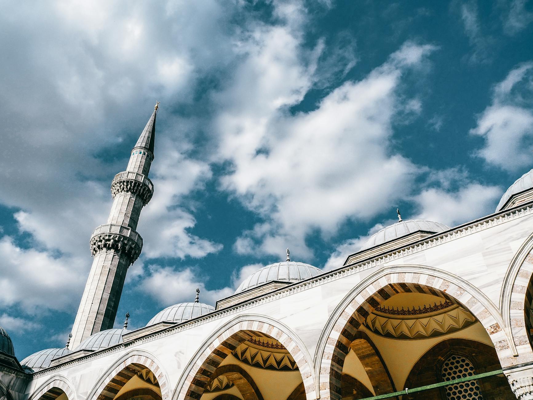 Low-angle shot of a soaring minaret and white marble arches of an Ottoman mosque against a dramatic blue and white cloudy sky, characteristic of Istanbul tourism.