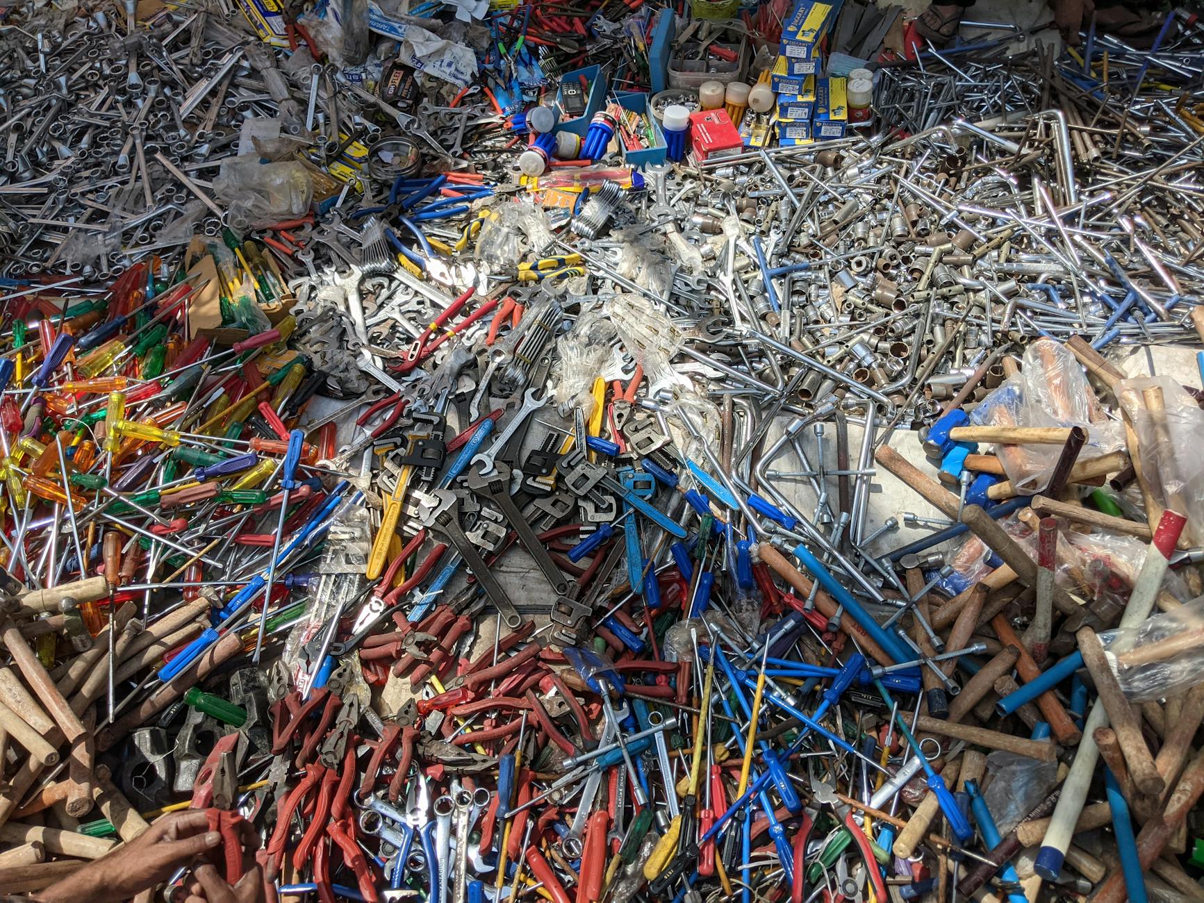 An overhead, vibrant close-up view of a chaotic pile of assorted metal tools, showcasing 'The Grease and Iron of My Favorite Walk Through Perşembe Pazarı.' The collection includes numerous shiny wrenches, pliers with red and blue handles, screwdrivers with colorful translucent grips, and various wooden-handled implements, typical of a bustling hardware section in a Turkish bazaar.