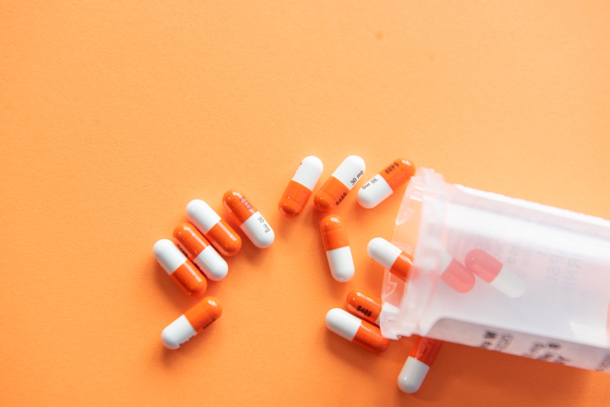 Organized pharmacy medicine capsules spilling from a prescription bottle on an orange background.