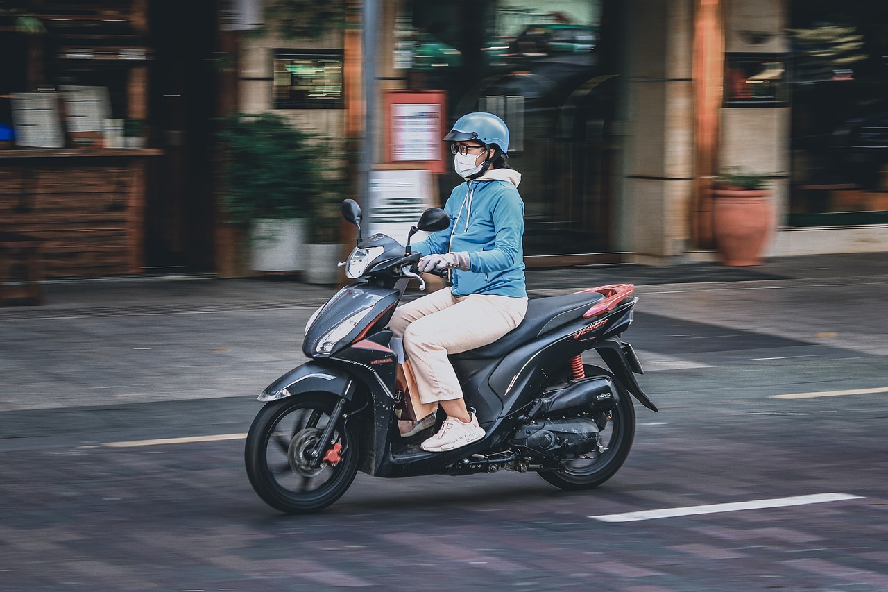 A person wearing a light blue hoodie, a helmet, and a face mask rides a dark-colored Honda scooter through city streets, illustrating the challenge of traffic. This dynamic shot suggests the hustle required, perhaps referencing 'The Only Reason I Brave the Traffic to Spend an Afternoon in Teşvikiye', although the specific location cannot be confirmed.