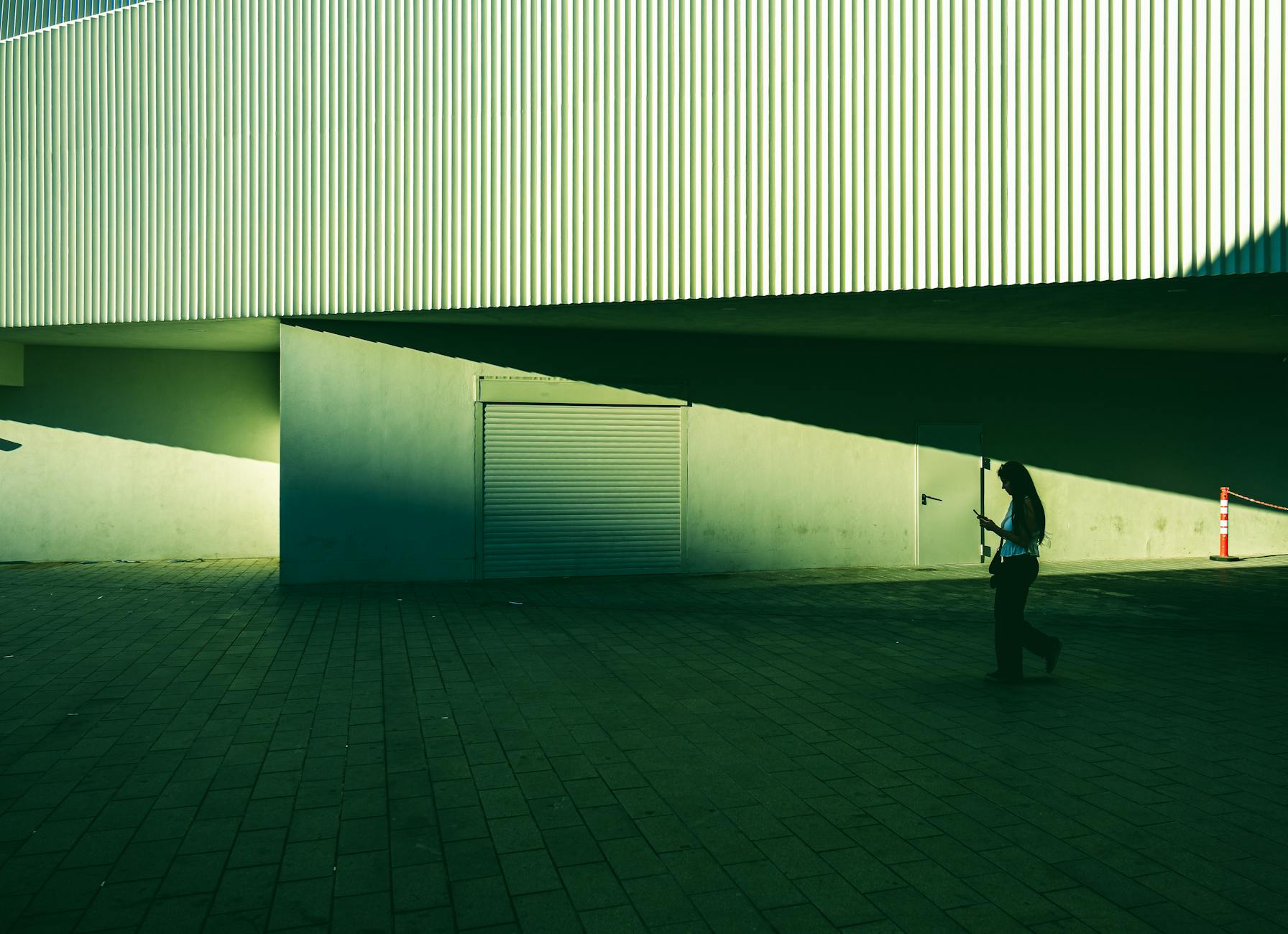 A dramatic, high-contrast street photograph featuring strong architectural lines and deep shadows, tinted green. A silhouetted woman walks across a tiled pavement while looking at her phone, embodying the gritty, lived-in feel suggested by the topic: 'Kurtuluş is loud and the sidewalks are broken but you should stay there anyway.' The strong light and shadow contrast highlight the texture of the modern building facade and the slightly uneven ground.