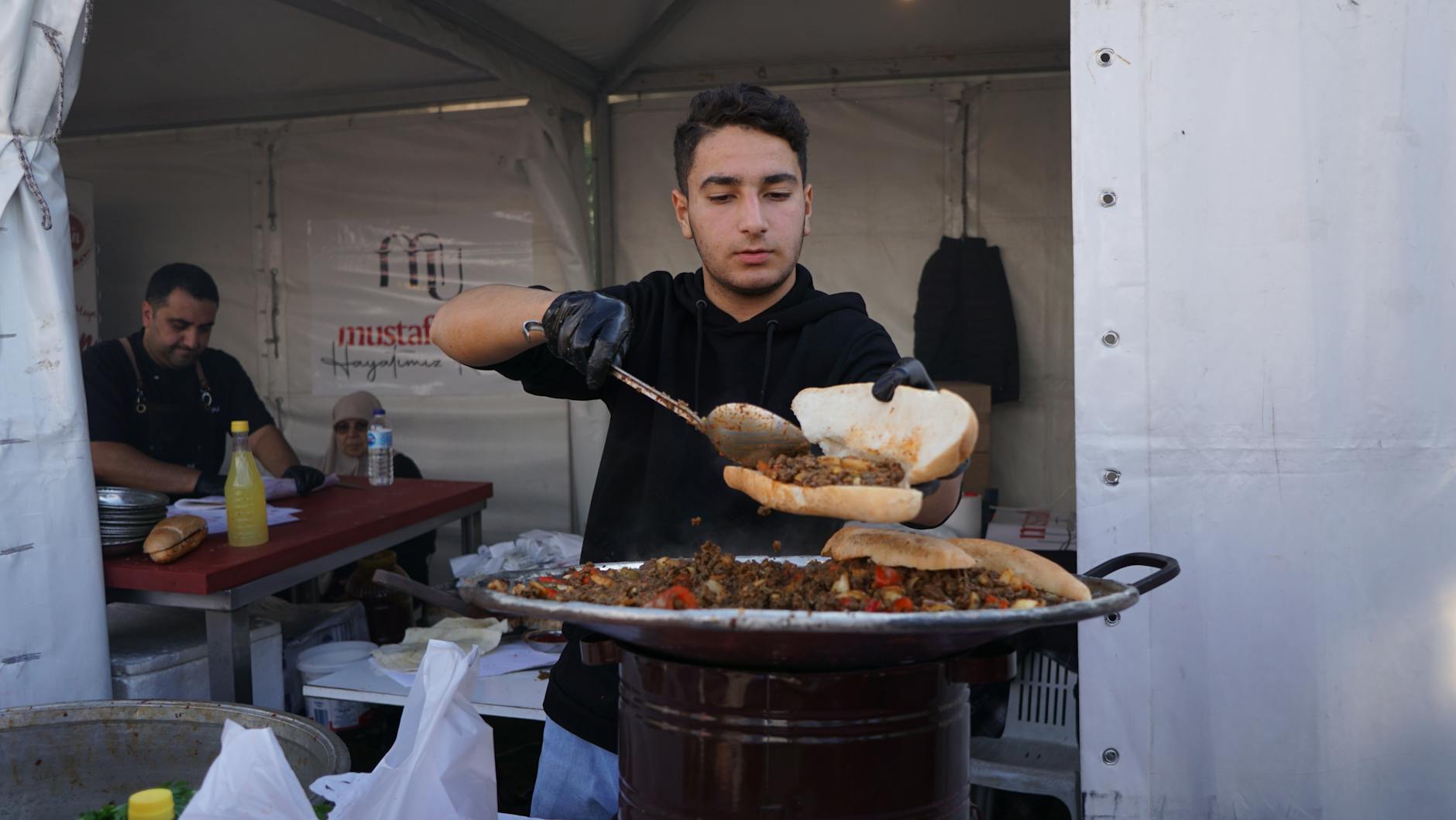A young chef prepares spicy Mersin style tantuni meat in a large circular metal pan.