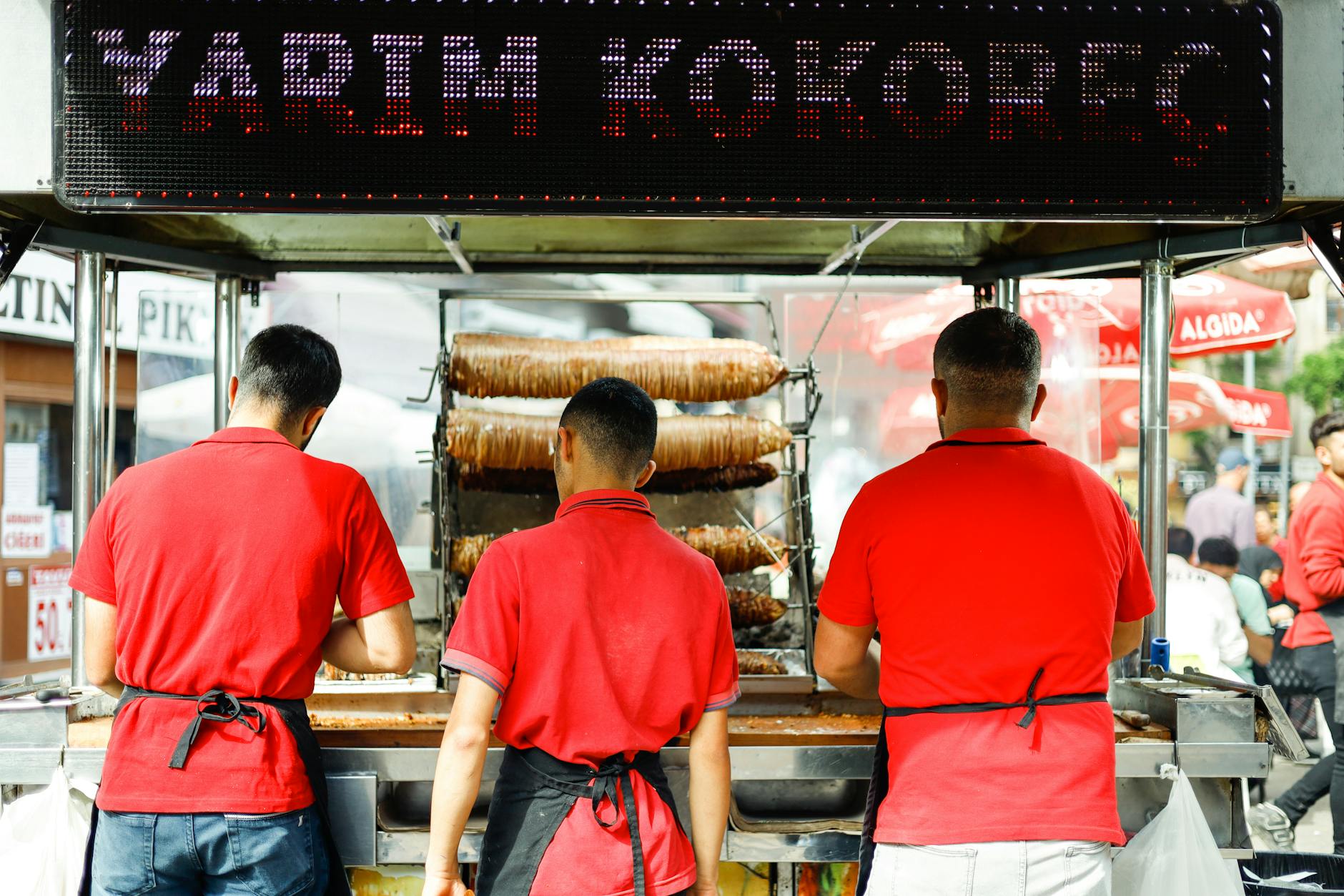 Turkish street food vendors stand before large spits of roasting meat under a bright sign.