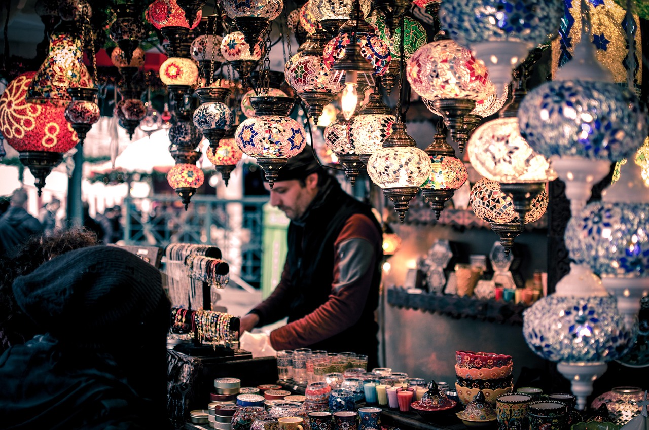 A vibrant scene under a canopy of hanging, illuminated Turkish mosaic lamps, characteristic of Istanbul markets. A vendor, blurred in the background, attends to his stall, which features bracelets, small candles, and colorful ceramic items. This image captures the atmospheric chaos that defines the experience of The frantic sweaty madness of my walk through Mahmutpaşa and Tahtakale.