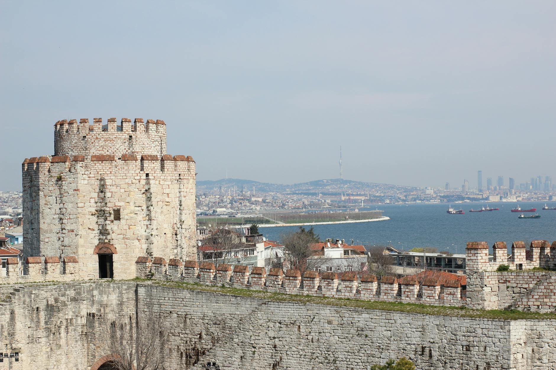 A panoramic view of the imposing stone walls and a circular tower of the Yedikule Fortress (Seven Towers) in Istanbul, Turkey. The ancient battlements stand prominently in the foreground, overlooking the sparkling blue waters of the Sea of Marmara dotted with cargo ships. In the hazy distance across the water, the modern skyline of the Asian side of Istanbul is visible, making the fortress feel like the only honest place left in the city, as mentioned in the article topic.