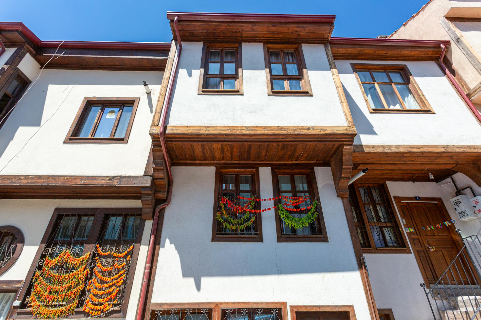 Low-angle view of a traditional Ottoman-style house facade in Istanbul's Zeyrek neighborhood, featuring white plaster walls, rich dark wood framing, and colorful drying peppers hanging from the window grates.