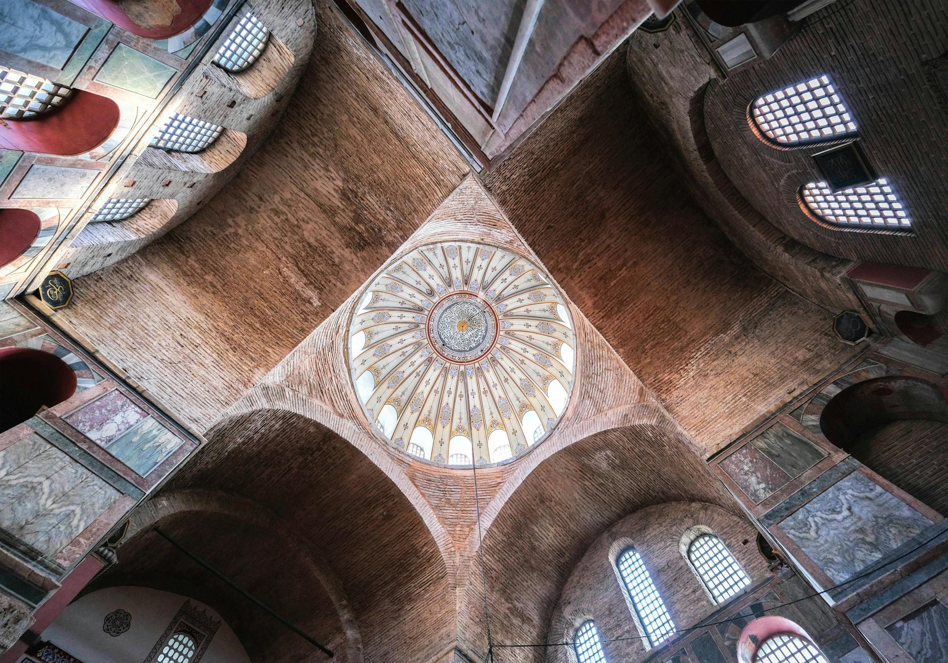 Dramatic low-angle view looking up at the intricate, light-filled dome and brick arches inside the historic Zeyrek Mosque in Istanbul, Turkey.